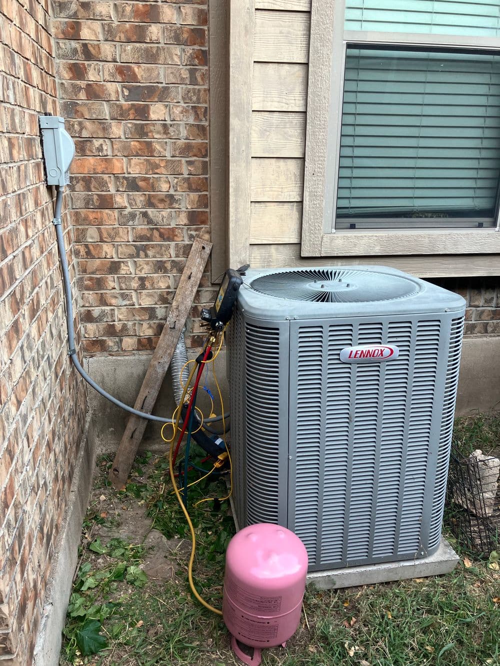 Lennox central air conditioning unit installed by a brick wall with visible wiring and pink tank.